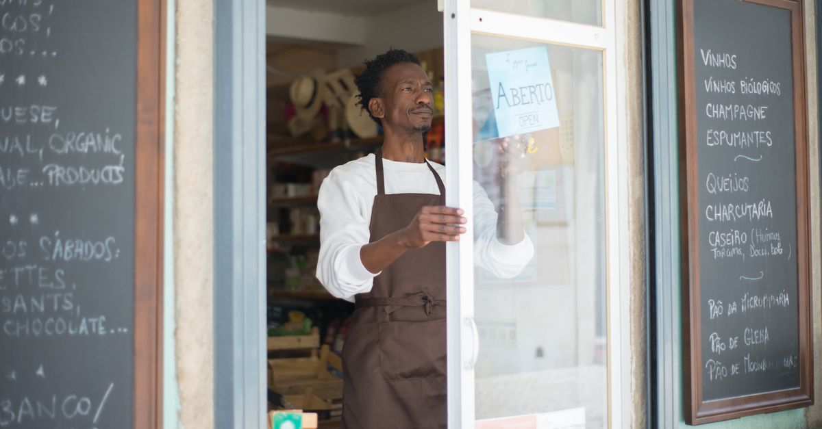 A man in an apron opens a grocery store door in Portugal, ready for business.